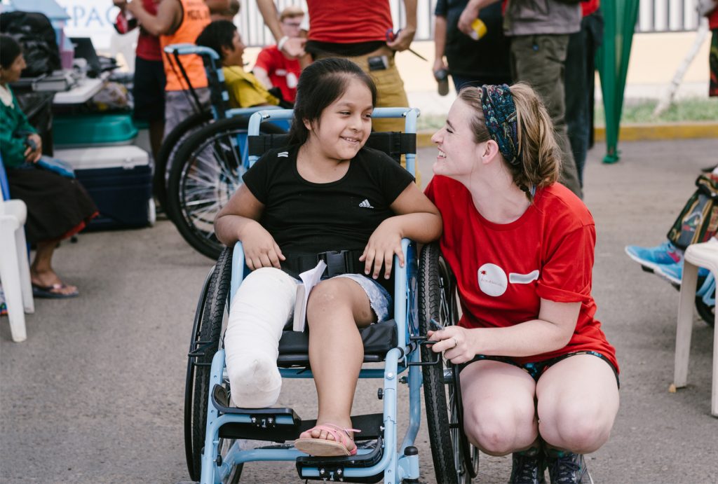 A smiling young woman kneels to meet a girl in a wheelchair eye-to-eye.