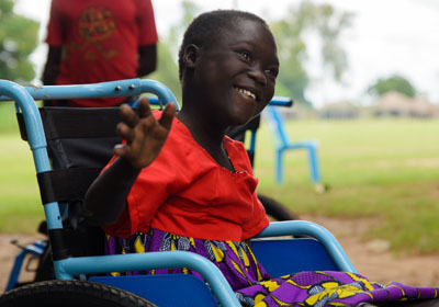 A smiling African girl in a red shirt sits in a new, blue wheelchair.