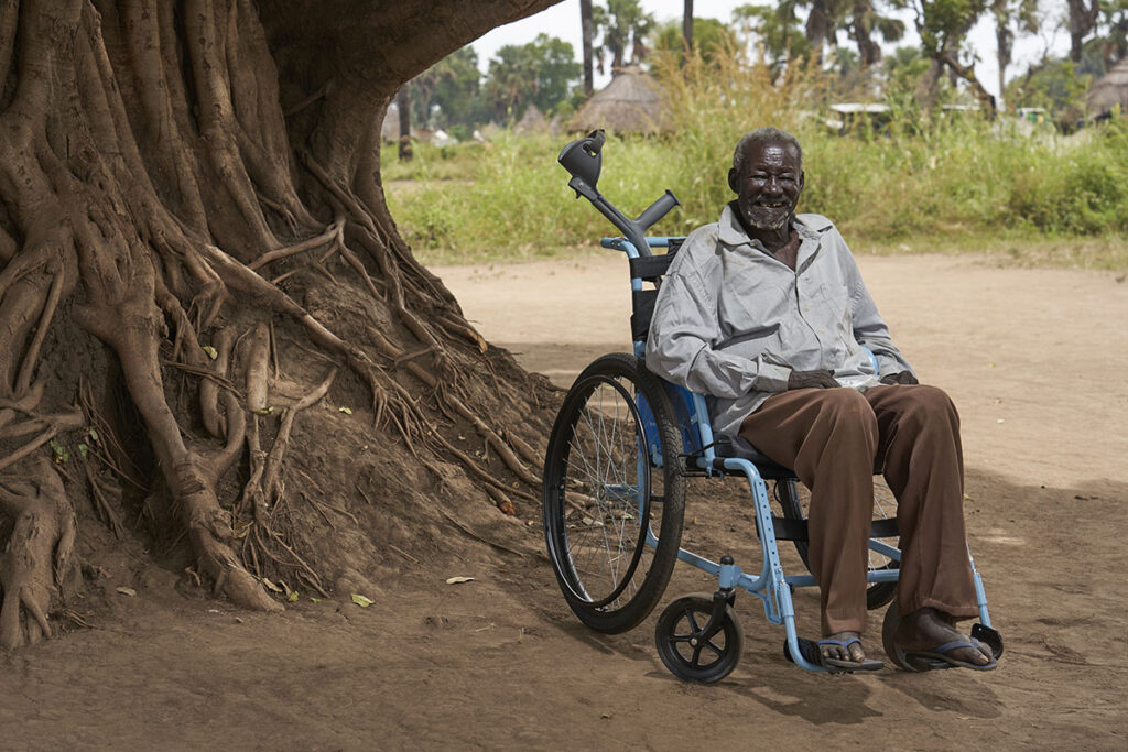 A man in a wheelchair, under a tree in Kenya, with his crutches in the back.