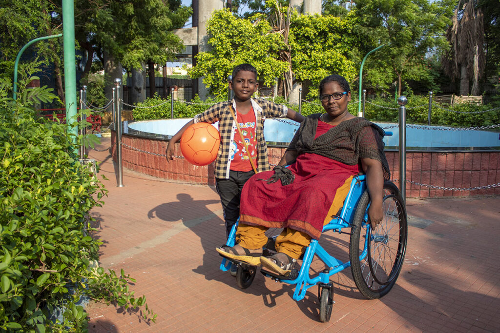 A boy holding a ball stands next to his mother, who is in a wheelchair, in India.
