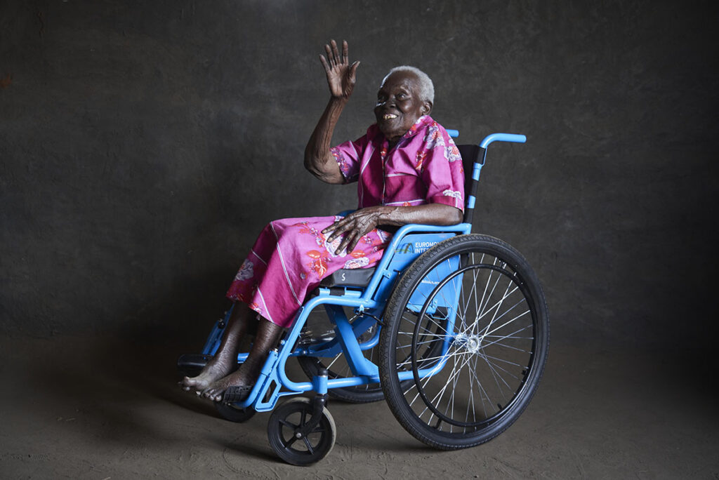 A woman in Kenya sits in a wheelchair and waves with a smile.
