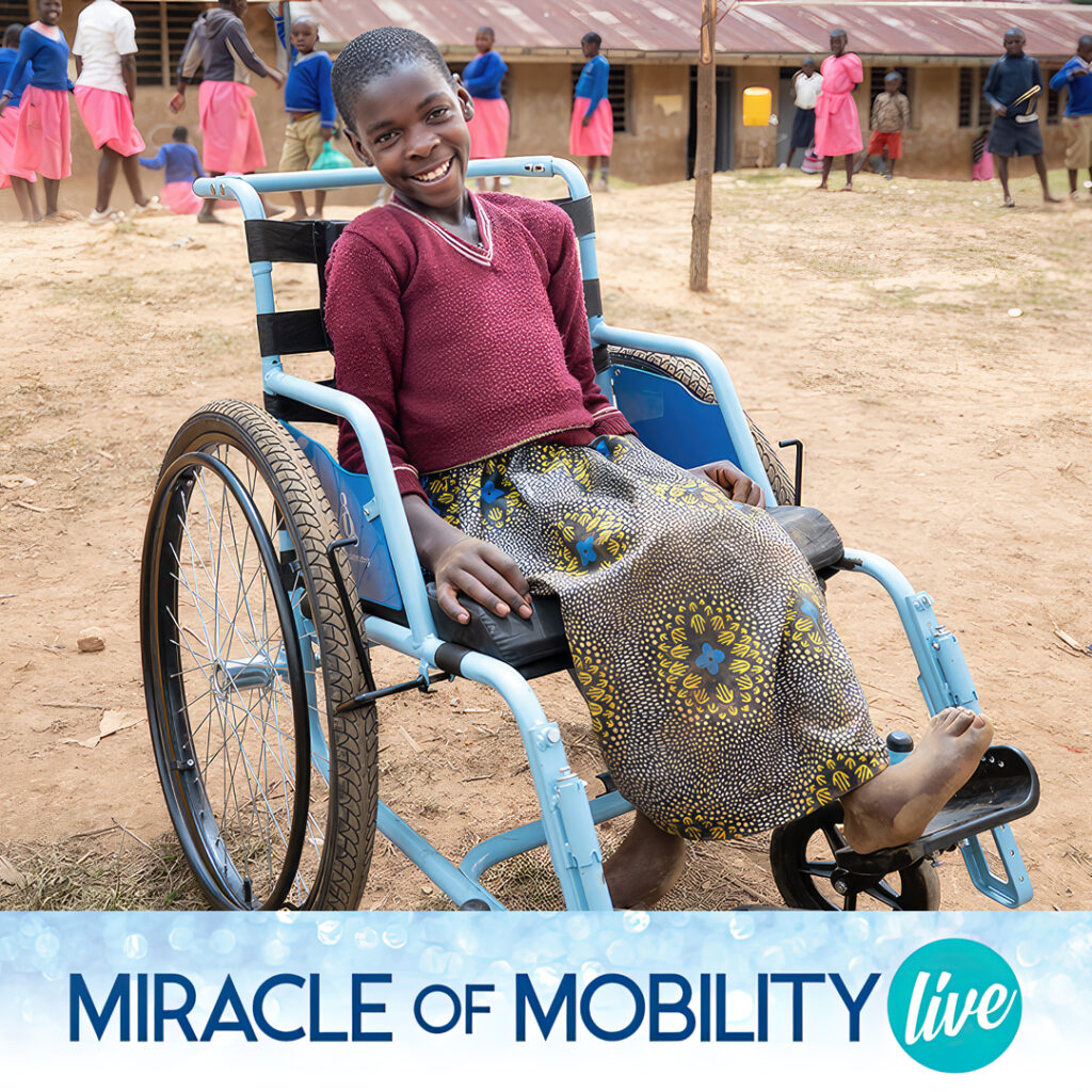 A smiling young woman in a wheelchair, on a field with the reddish soil of Uganda.