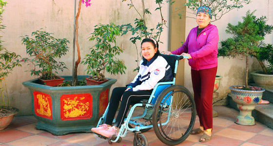 A Vietnamese girl named Anh in a wheelchair, smiling, being pushed past some potted plants by her mother.