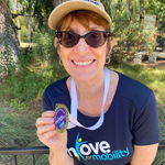 A smiling woman wearing sunglasses, holding up a medal.