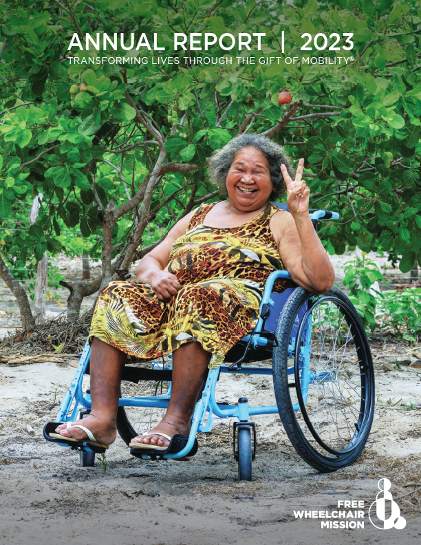 An indigenous woman in Brazil smiles, holding up the peace sign as she sits outdoors in a new blue wheelchair.